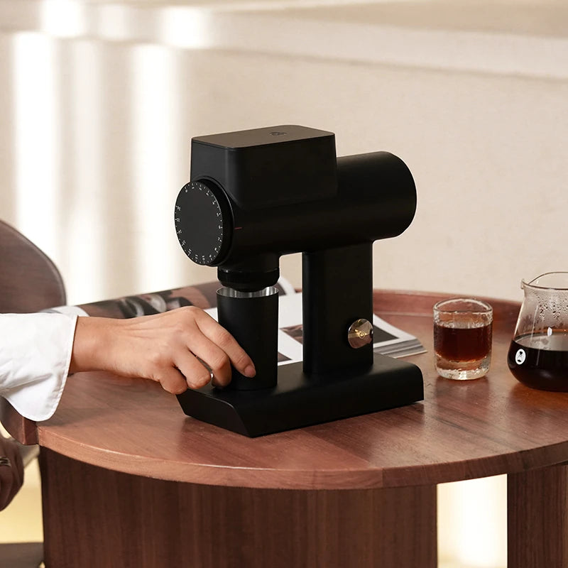 Timemore Sculptor black coffee grinder on a wooden table with a glass of coffee and a carafe in the background.