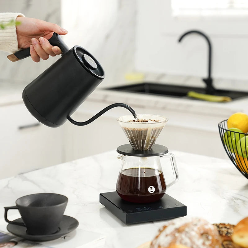 Person pouring coffee from a black kettle into a glass carafe on a kitchen counter.