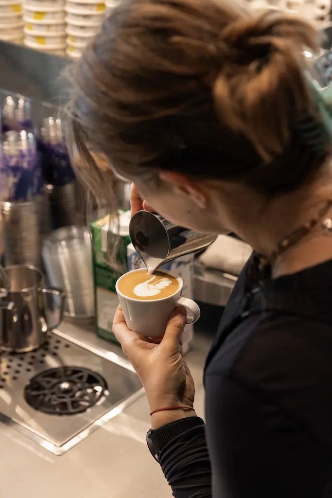Person pouring a spoonful of cream into a coffee cup in a kitchen setting.
